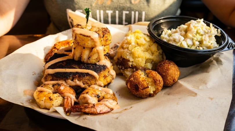 A seafood platter with grilled shrimp, scallops, mashed potatoes, hush puppies, and coleslaw.