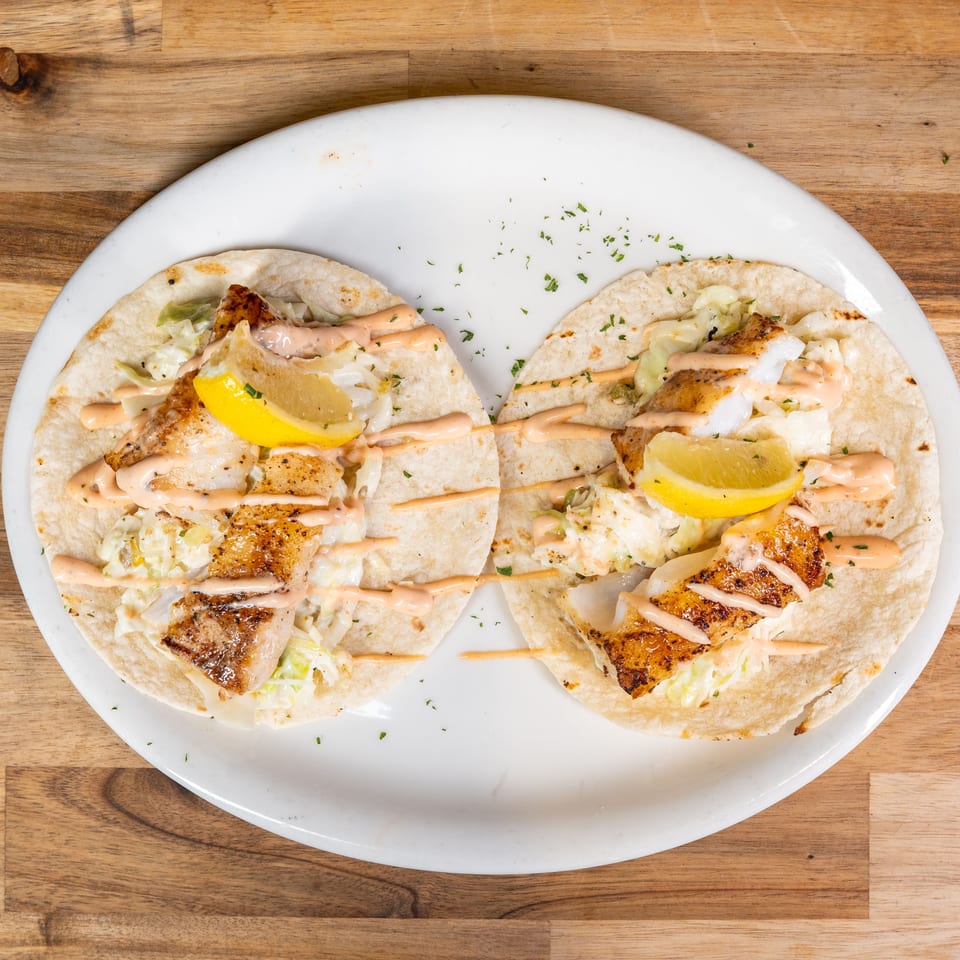 Two fish tacos with slaw, sauce, and lemon wedges on a white plate atop a wooden table.