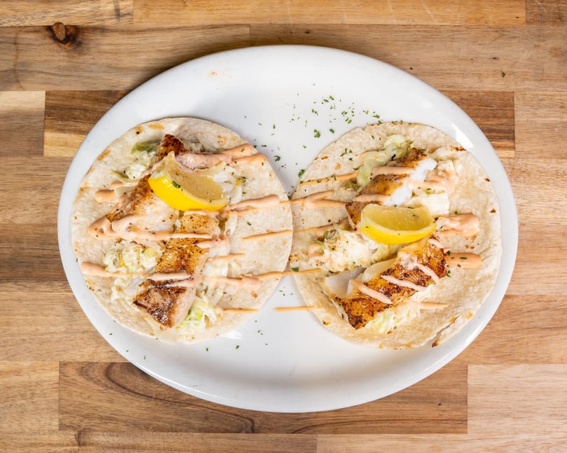 Two fish tacos with slaw, sauce, and lemon wedges on a white plate atop a wooden table.