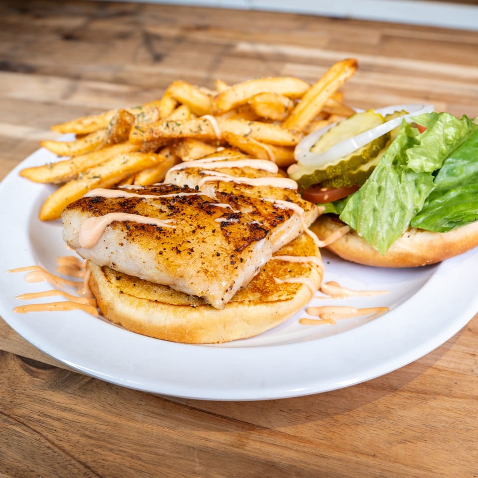 Grilled fish sandwich with fries, lettuce, tomato, pickles, and onions on a white plate.