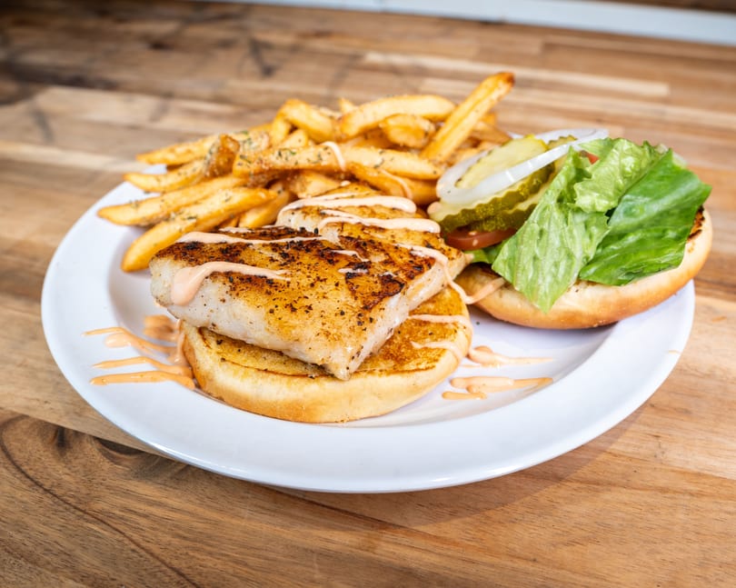 Grilled fish sandwich with fries, lettuce, tomato, pickles, and onions on a white plate.