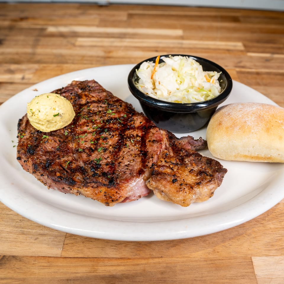 Grilled steak with a pat of butter, coleslaw, and a dinner roll on a white plate.