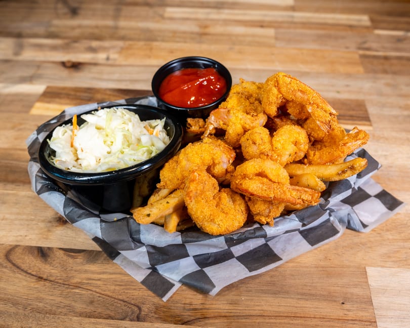 Fried shrimp and french fries in a basket with coleslaw and ketchup on a wooden table.