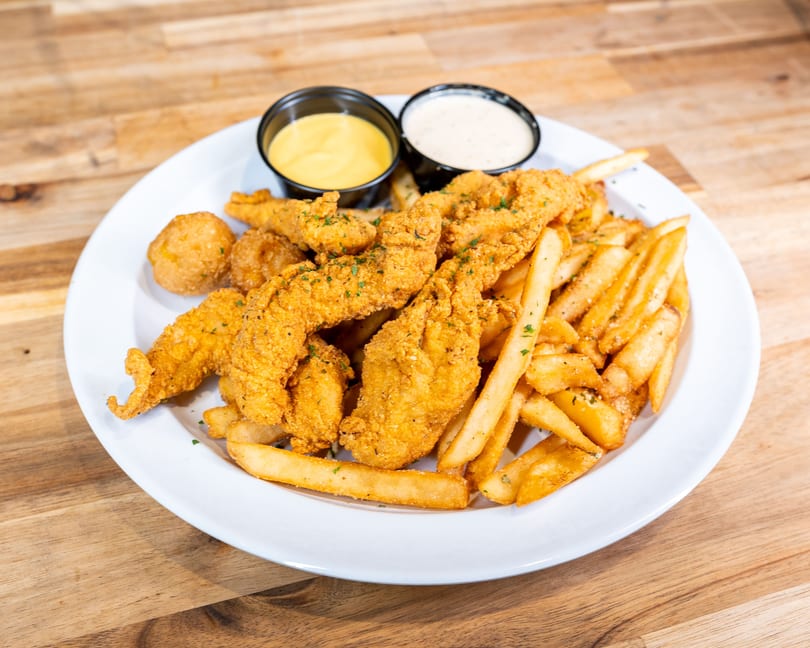 Crispy fried chicken tenders, hush puppies, and french fries with two dipping sauces on a white plate.